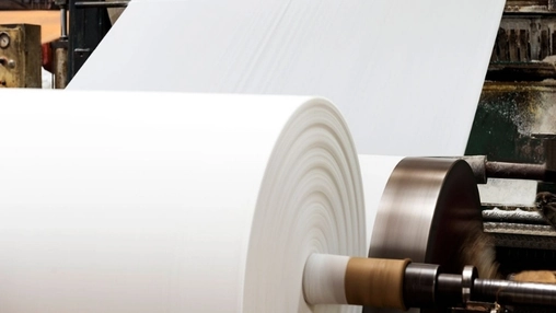 Industrial paper mill machine processing a large white paper roll with a steel drum and worn green machinery